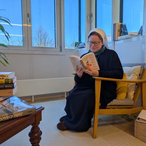 Eine Ordensschwester in Ordenskleidung sitzt an einem Fenster in einem Sessel und liest in einem Buch. Vor ihr ist ein Tisch mit einem Bücherstapel zu sehen.