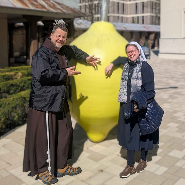 Ein Mönch und eine Schwester in Ordenskleidung stehen rechts und links von einer großen Zitrone und blicken freundlich in die Kamera.